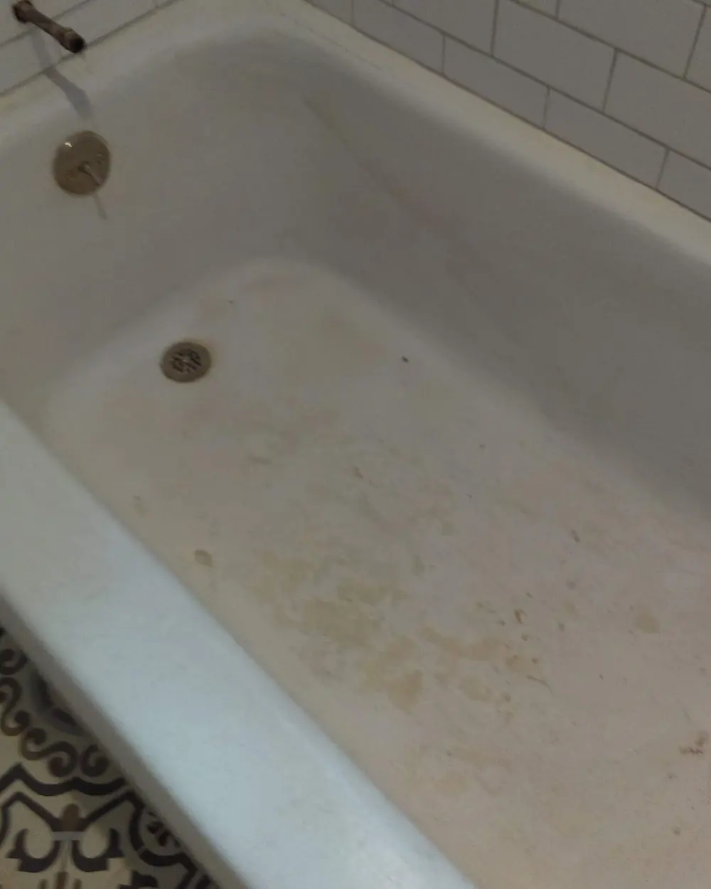 A cast-iron bathtub before refinishing, the basin yellowed and stained brown across the floor and around the drain, against a white subway-tile wall.